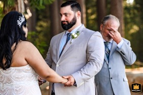 In Lake Tahoe, California, the emotional father of the groom stands behind his son during the ceremony, quietly wiping away a single tear in a powerful moment of pride and love.